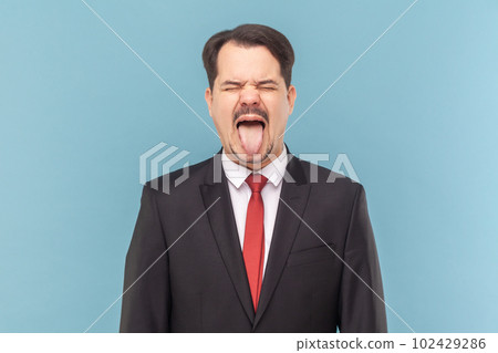 Portrait of crazy excited man with mustache standing sticking tongue out, demonstrates childish behavior, wearing black suit with red tie. Indoor studio shot isolated on light blue background. 102429286