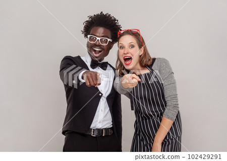 Portrait of excited happy positive woman and man in white glasses standing pointing at you, laughing out loud, shame on you. Indoor studio shot isolated on gray background. 102429291