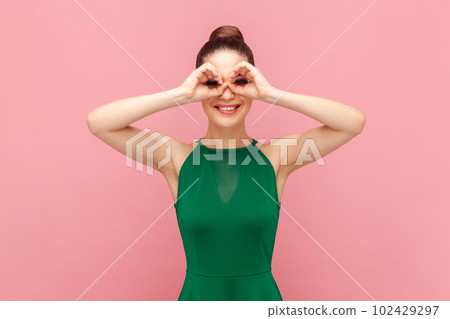 Portrait of positive smiling happy woman looking at camera, imagines she looks through binoculars, sees something funny, wearing green dress. Indoor studio shot isolated on pink background. 102429297