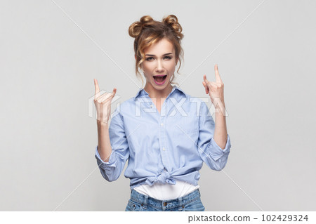 Portrait of blonde woman showing rock and roll gesture heavy metal sign enjoying favorite music on party has fun, exclaims from joy, wearing blue shirt. Indoor studio shot isolated on gray background. 102429324