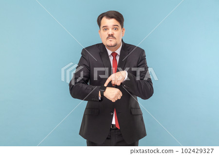 Portrait of serious man with mustache standing looking at camera, pointing at his smartwatch, deadline, wearing black suit with red tie. Indoor studio shot isolated on light blue background. 102429327