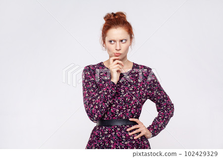 Portrait of redhead woman wearing dress holding chin and pondering idea, planning strategy, musing and solving difficult question in mind. Indoor studio shot isolated on gray background. 102429329