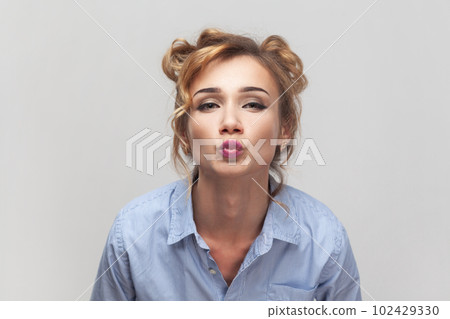 Air kiss for you. Portrait of blonde woman keeps lips folded, expressing love, blowing mwah, having romantic expression, wearing blue shirt. Indoor studio shot isolated on gray background. Air kiss for you. Portrait of blonde woman keeps lips folded, expressing love, blowing mwah, having romantic expression, wearing blue shirt. Indoor studio shot isolated on gray background. 102429330