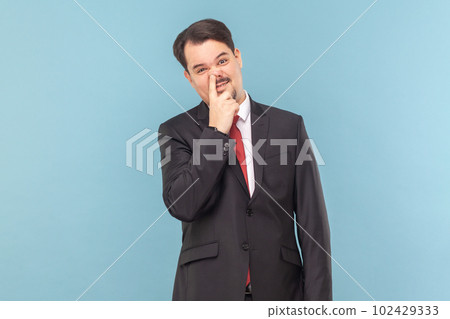 Portrait of funny childish man with mustache standing having comical facial expression, keeps finger in nose, wearing black suit with red tie. Indoor studio shot isolated on light blue background. 102429333