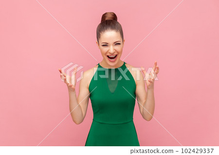 Portrait of crazy angry woman standing with raised arms, screaming with aggression, having problems, shouting, wearing green dress. Indoor studio shot isolated on pink background. 102429337