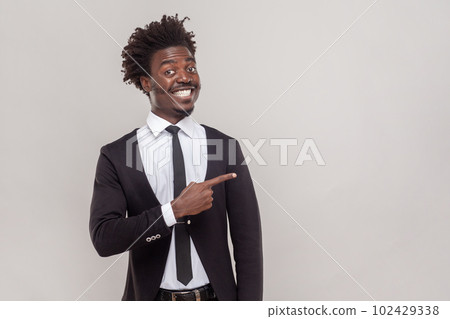 Man with Afro hairstyle has happy expression, smiles gently, points aside with fore finger, shows place to hang out, wearing white shirt and tuxedo. Indoor studio shot isolated on gray background. 102429338