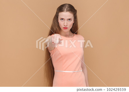 Portrait of serious strict woman with long hair standing pointing to camera, choosing you, select, making choice, wearing elegant dress. Indoor studio shot isolated on brown background. 102429339