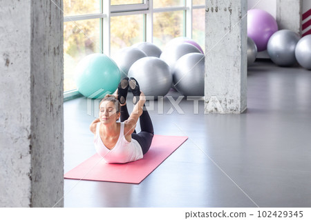 Portrait of sporty and flexible woman lying on bellies and holding legs in hands while practicing yoga on exercise mat, keeps eyes closed. Indoor shot with window on background. 102429345