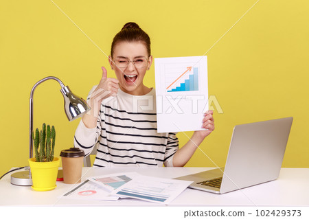 Delighted woman smiling at camera holding growth diagram, showing thumb up, satisfied with financial and economic growth of his business. Indoor studio studio shot isolated on yellow background. Delighted woman smiling at camera holding growth diagram, showing thumb up, satisfied with financial and economic growth of his business. Indoor studio studio shot isolated on yellow background. 102429373