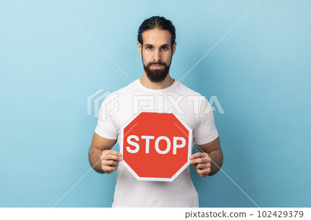 Portrait of serious man with beard wearing white T-shirt holding red Stop sign, looking at camera with negative aggressive expression, showing ban. Indoor studio shot isolated on blue background. Portrait of serious man with beard wearing white T-shirt holding red Stop sign, looking at camera with negative aggressive expression, showing ban. Indoor studio shot isolated on blue background. 102429379