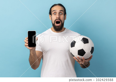 Portrait of amazed man with beard wearing white T-shirt holding soccer ball and smartphone empty black display, ticket booking for championship. Indoor studio shot isolated on blue background. 102429388