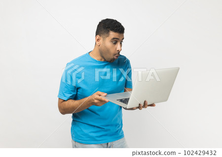 Portrait of surprised astonished unshaven man wearing blue T- shirt standing holding laptop in hands, working online, looking at screen with big eyes. Indoor studio shot isolated on gray background. 102429432