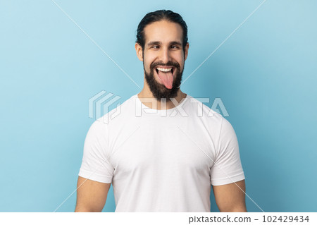 Portrait of comic positive man with beard wearing white T-shirt looking cross-eyed, having fun with silly face expression, showing tongue out. Indoor studio shot isolated on blue background. 102429434