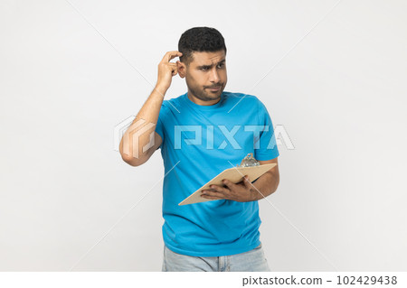 Portrait of pensive puzzled unshaven man wearing blue T- shirt standing with clipboard, writing something on paper, thinking about plan. Indoor studio shot isolated on gray background. 102429438