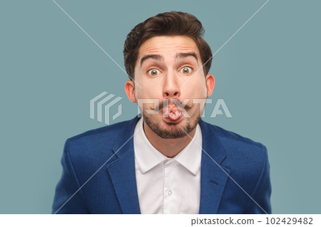 Portrait of funny handsome man with mustache standing sticking tongue out, showing childish behavior, wearing white shirt and jacket. Indoor studio shot isolated on light blue background. 102429482