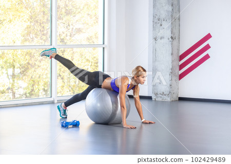 Side view portrait of slim attractive sporty woman lying on a fitness ball with her palms on floor, raised her leg up, wearing sports top and tights. Indoor shot with window on background. 102429489