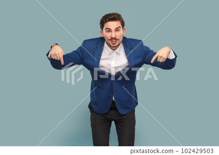 Angry man with mustache standing pointing down, giving command, looking at camera with serious expression, wearing white shirt and jacket. Indoor studio shot isolated on light blue background. 102429503