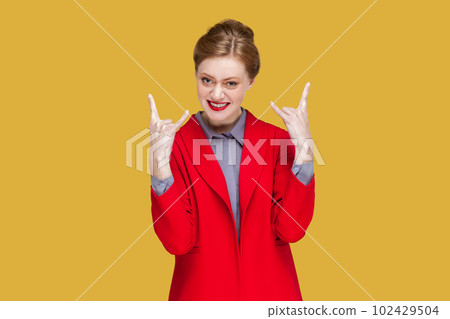Portrait of excited crazy woman with red lips standing showing rock and roll gesture, having fun on festival, wearing red jacket. Indoor studio shot isolated on yellow background. Portrait of excited crazy woman with red lips standing showing rock and roll gesture, having fun on festival, wearing red jacket. Indoor studio shot isolated on yellow background. 102429504