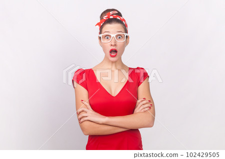 Portrait of shocked surprised woman wearing red dress, glasses and head band standing with crossed arms, looking at camera with big eyes and open mouth. Indoor studio shot isolated on gray background. 102429505
