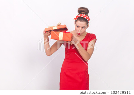 Portrait of curious attractive woman wearing red dress and head band holding present box, looking inside with interest, having gift on her birthday. Indoor studio shot isolated on gray background. Portrait of curious attractive woman wearing red dress and head band holding present box, looking inside with interest, having gift on her birthday. Indoor studio shot isolated on gray background. 102429511