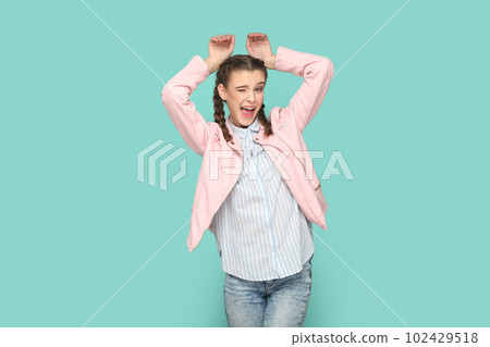 Portrait of funny positive childish teenager girl with braids wearing pink jacket showing bunny ears, winking to camera. Indoor studio shot isolated on green background. 102429518