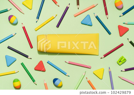 Yellow pencil case in centre with Colored Pencils and rubber erasers background pattern on light green. Back to School or drawing and creativity concept. Copy space in center. Top view 102429879