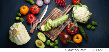 Fresh various vegetables, cut zucchini on wooden cutting board and knife on rustic dark background top view. Cooking vegetarian meal with healthy ingredients, diet food and nutrition concept Fresh various vegetables, cut zucchini on wooden cutting board and knife on rustic dark background top view. Cooking vegetarian meal with healthy ingredients, diet food and nutrition concept 102432733