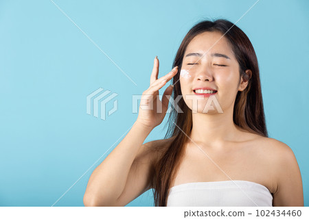 Happy beauty portrait woman gets cream on her cheek, Beautiful young female smiling applying cosmetic cream on her face isolated on blue background, health clean skin, Skin care and beauty concept Happy beauty portrait woman gets cream on her cheek, Beautiful young female smiling applying cosmetic cream on her face isolated on blue background, health clean skin, Skin care and beauty concept 102434460