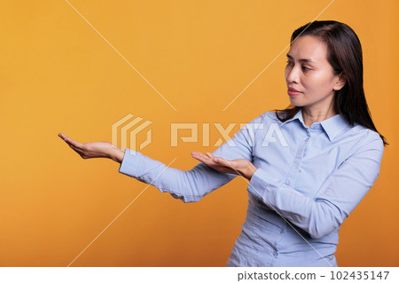 Filipino model pointing at presentation icon on camera, doing advertisement and showing example product. Cheerful woman posing over yellow background in studio showing confident girl doing ad. 102435147