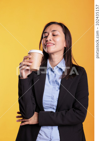Satisfied asian model holding cup of coffee drinking hot beverage enjoying free time before start to photoshot in studio. cheerful young adult with brunette long hair standing over yellow background 102435157