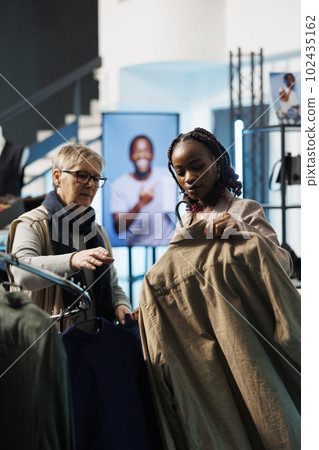 African american employee helping elderly client choosing brown shirt for husband in modern boutique. Senior woman shopping for casual wear, buying fashionable merchandise in clothing store 102435162