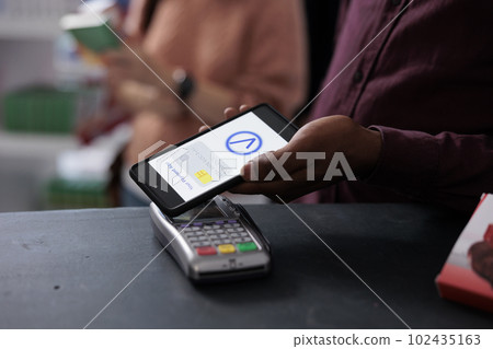 Young adult putting phone on pos terminal using contactless to pay for pharmaceutics medication in pharmacy, shopping for pain pills. Clients waiting in line at drugstore counter. Close up Young adult putting phone on pos terminal using contactless to pay for pharmaceutics medication in pharmacy, shopping for pain pills. Clients waiting in line at drugstore counter. Close up 102435163