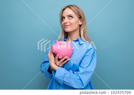 dreaming blond young adult hugging a piggy bank where she keeps savings on a blue isolated dreaming blond young adult hugging a piggy bank where she keeps savings on a blue isolated 102435709