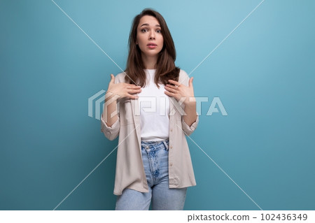 surprised puzzled 30s dark haired woman in shirt and jeans on blue background 102436349
