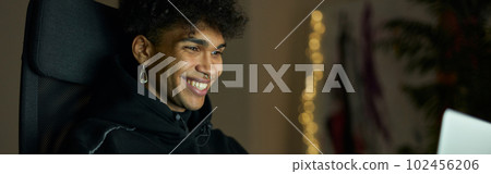 Cheerful young guy with piercing smiling while sitting in front of laptop screen late in the evening Cheerful young guy with piercing smiling while sitting in front of laptop screen late in the evening 102456206