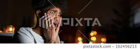 Young guy looking terrified while watching horror movie alone using laptop and eating popcorn, sitting on a comfortable sofa in dark room at home 102456213