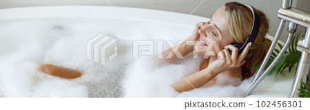 Cheerful young woman in headphones smiling, listening to music in bath with bubbles. 102456301