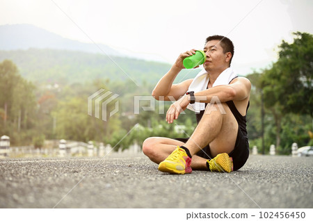Sweaty and thirsty mature Asian man sits on the road in a public park and drinks water Sweaty and thirsty mature Asian man sits on the road in a public park and drinks water 102456450