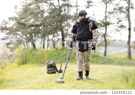 A man in a military uniform and bulletproof vest works in the forest with a metal detector. A minesweeper performs work on demining the territory A man in a military uniform and bulletproof vest works in the forest with a metal detector. A minesweeper performs work on demining the territory 102458122