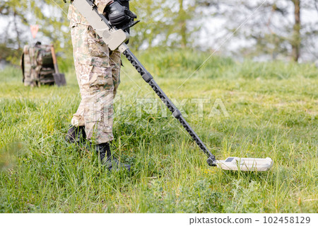 A man in a military uniform and bulletproof vest works in the forest with a metal detector. A minesweeper performs work on demining the territory A man in a military uniform and bulletproof vest works in the forest with a metal detector. A minesweeper performs work on demining the territory 102458129