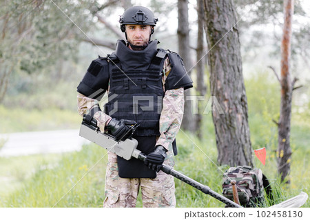 A man in a military uniform and bulletproof vest works in the forest with a metal detector. A minesweeper performs work on demining the territory A man in a military uniform and bulletproof vest works in the forest with a metal detector. A minesweeper performs work on demining the territory 102458130