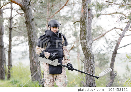 A man in a military uniform and bulletproof vest works in the forest with a metal detector. A minesweeper performs work on demining the territory 102458131