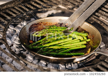 Close-up of green frying asparagus outdoor on BBQ grill. Cocking vegetables on carbon steel pan. Using kitchen tongs. Hot coals 102458260
