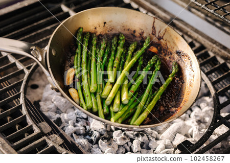 Delicious preparation of green asparagus cooked in butter with garlic in a frying pan on outdoor BBQ grill.  102458267