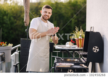 Portrait of handsome young chief in apron stand by the BBQ grill and smile. Male look into camera and rubs hands. Portrait of handsome young chief in apron stand by the BBQ grill and smile. Male look into camera and rubs hands. 102458268