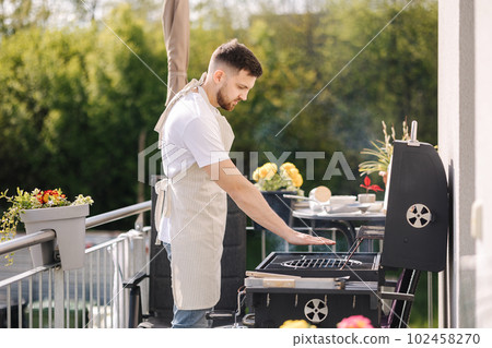 Side view of handsome man try how hot is BBQ grill. Man in apron make dinner outdoor 102458270