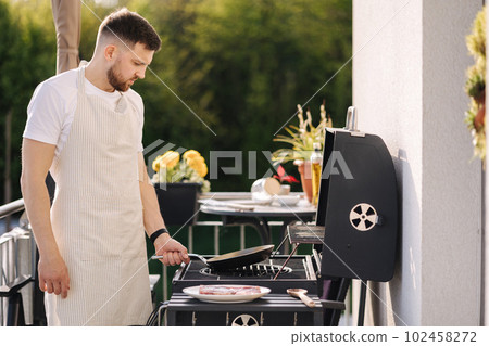Handsome bearded man in apron frying potatoes on BBQ grill using frying carbon steel pan.  102458272