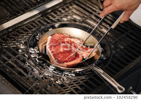 Human hand using grilling tongs to flip the steak on pan. Grill outdoor.  102458290