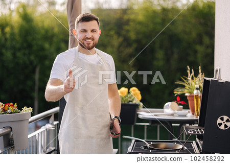 Portrait of handsome young chief in apron stand by the BBQ grill and smile. Male look into camera and showing thumbs up. 102458292