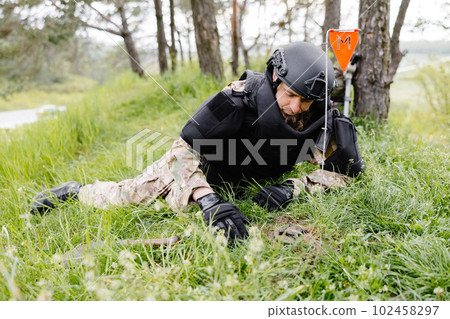A man in a military uniform and a bulletproof vest works in the forest to demine the territory. A man warns of danger by making a red mark 102458297